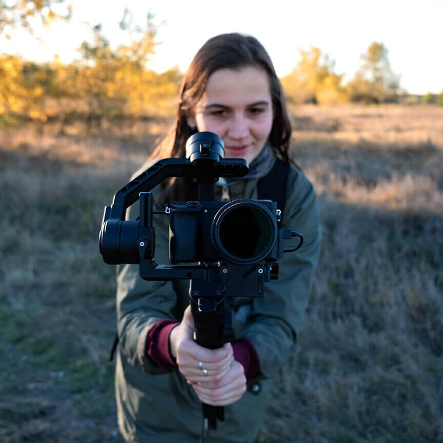 Female videographer holding a gimbal with mirrorless camera. Woman with stabilized camera rig filming outdoors on a sunny afternoon